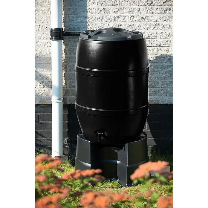 Nature Regentonnensockel - Länge: 53 cm, Schwarz - Eine Regentonne aus Kunststoff in Schwarz, die auf einem schwarzen Sockel steht. Ein Fallrohr ist an einer Ziegelwand befestigt.