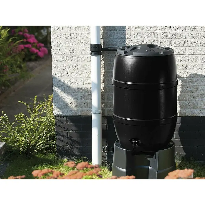 Nature Regentonnensockel - Länge: 53 cm, Schwarz - Ein schwarzer Wasserbehälter steht auf einem grauen Sockel vor einer weißen Ziegelwand und einem weißen Fallrohr.