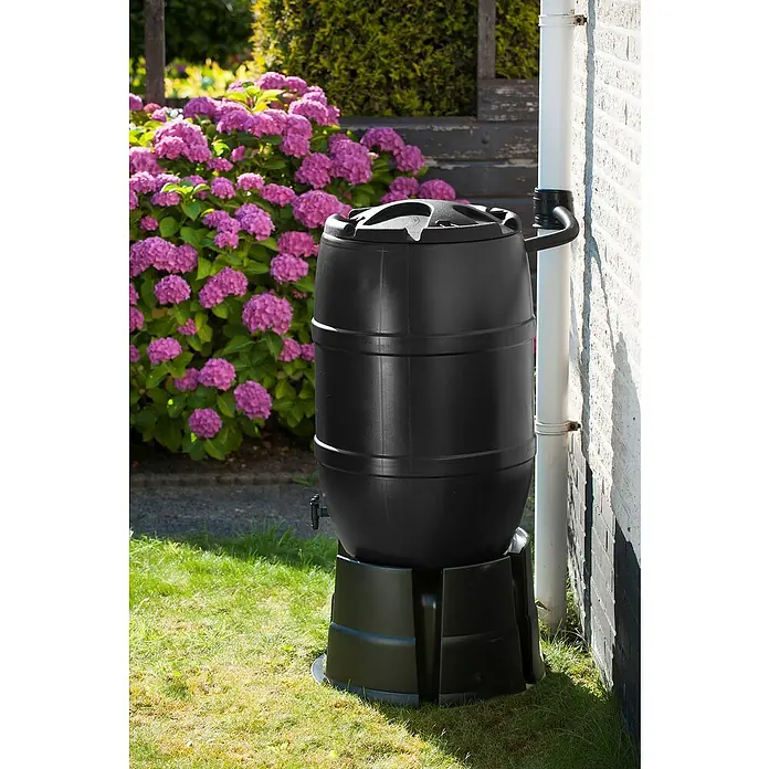 Nature Regentonne - 120 l, Grün - Eine schwarze Regentonne mit Deckel und Ständer, die neben einem weißen Fallrohr an einer Ziegelwand steht.