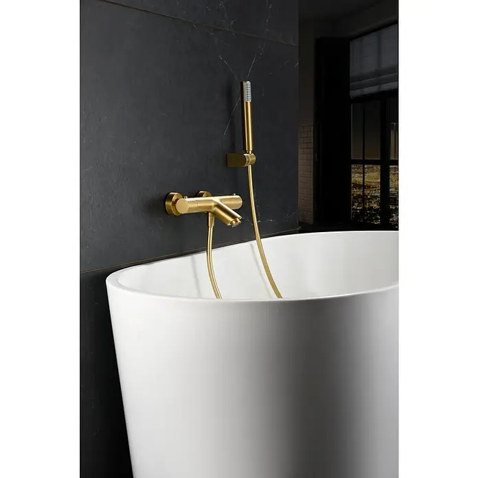 Imex Génova Grifo monomando de bañera - oro - Bañera moderna con grifería dorada y ducha de mano contra una pared oscura. Diseño elegante para una experiencia de baño lujosa.