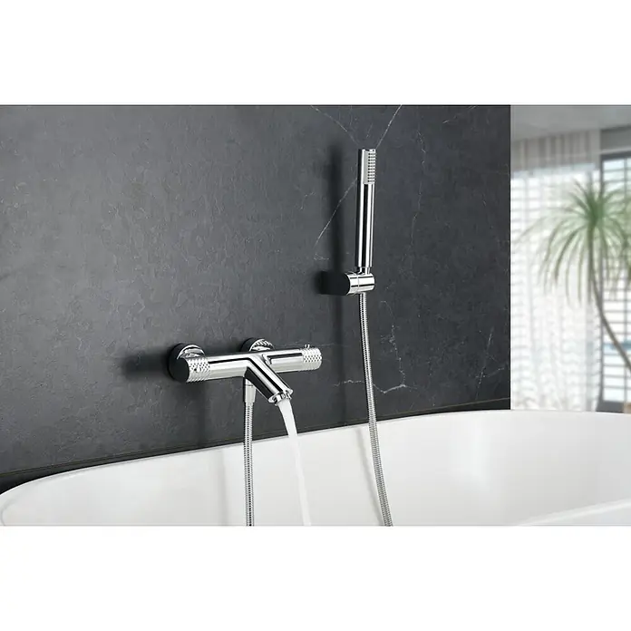 Imex Génova Grifo monomando de bañera - Cromo, Resplandeciente - Un moderno grifo de bañera cromado con teleducha y agua corriente, montado en una pared oscura y texturizada sobre una bañera blanca.