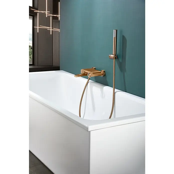 Imex Olimpo Grifo monomando de bañera - Oro rosa - Primer plano de una bañera con grifería dorada y alcachofa de ducha contra una pared color verde azulado.
