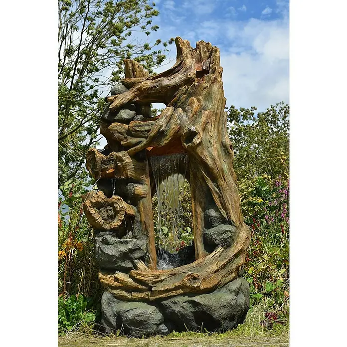 Ubbink AcquaArte Standbrunnen Kalloni - 207 x 104 x 104 cm - Nahaufnahme eines dekorativen Baumstamm-Brunnens mit fließendem Wasser, umgeben von üppigem Grün und einem bewölkten Himmel im Hintergrund.
