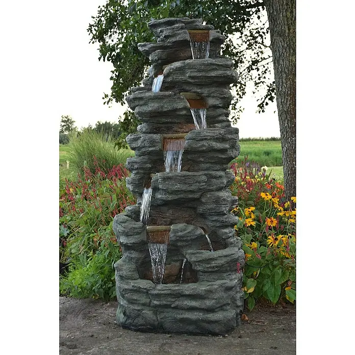 Ubbink AcquaArte Standbrunnen Corte - 70 x 87 x 201 cm - Ein mehrstufiger Gartenbrunnen aus grauem Kunststein mit fließendem Wasser inmitten blühender Pflanzen sorgt für eine entspannende Atmosphäre.