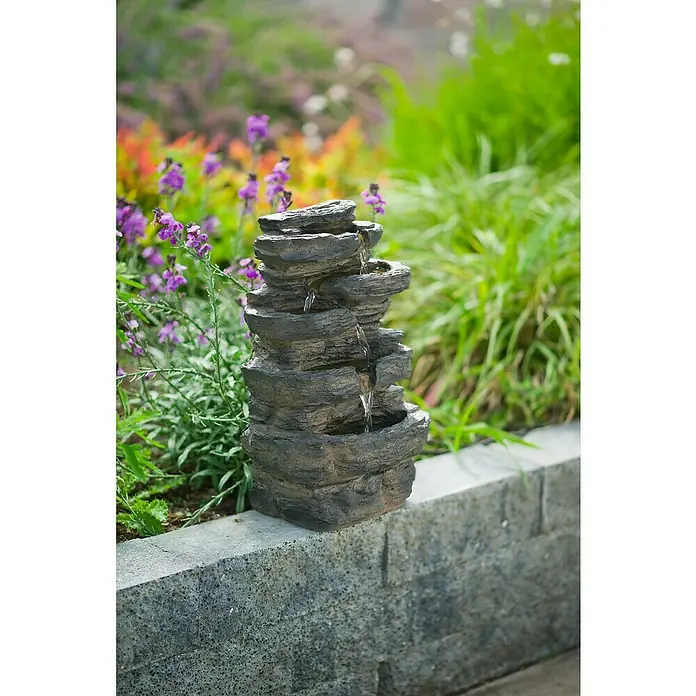 Ubbink AcquaArte Standbrunnen - 26 x 20 x 42 cm - Nahaufnahme eines mehrstufigen Zimmerbrunnens im Freien, umgeben von üppigem Grün und bunten Blumen.