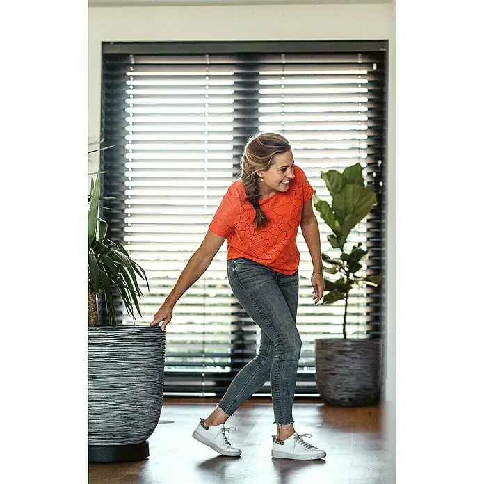 Capi Europe Nature Rib NL Pflanztopf Rib - Außenmaß (Ø x H): 43 x 41 cm, Kunststoff, Anthrazit - Frau schiebt Topfpflanze im Haus. Die Pflanze befindet sich in einem dekorativen, grau-melierten Topf mit horizontalem Linienmuster.