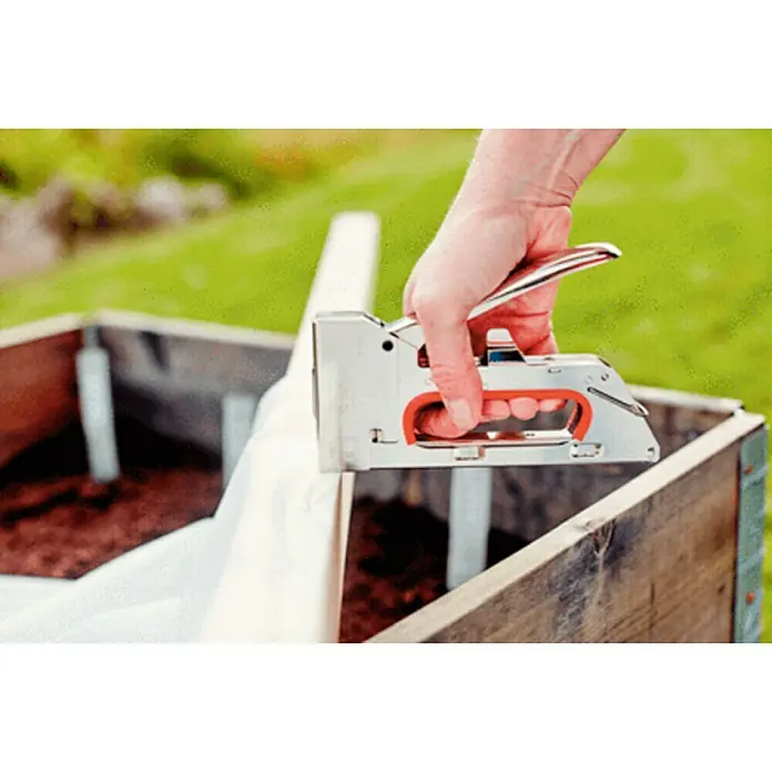 Rapid Grapadora manual 3153 - Profundidad de grapado: 4 mm - 8 mm - Un hombre está fijando una tela blanca a una caja de madera en el jardín con una grapadora. La grapadora es plateada con mango rojo.