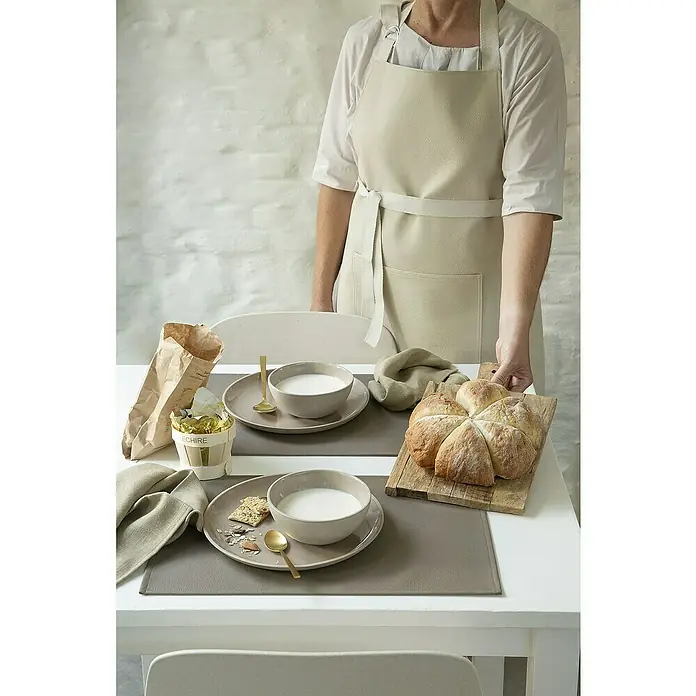 Tisch-Set Monaco - Taupe, 45 x 30 cm - Ein gedeckter Tisch mit grauen Platzsets, Schalen mit Milch, Brot und einer Person mit einer beigen Schürze im Hintergrund.