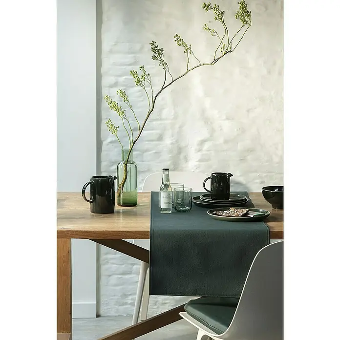 Tischläufer Monaco - 140 x 45 cm, Jungle Green - Elegante Tischdekoration mit dunklem Geschirr, Gläsern und einer schmalen Tischdecke auf einem Holztisch, dekoriert mit einem grünen Zweig in einer Vase.
