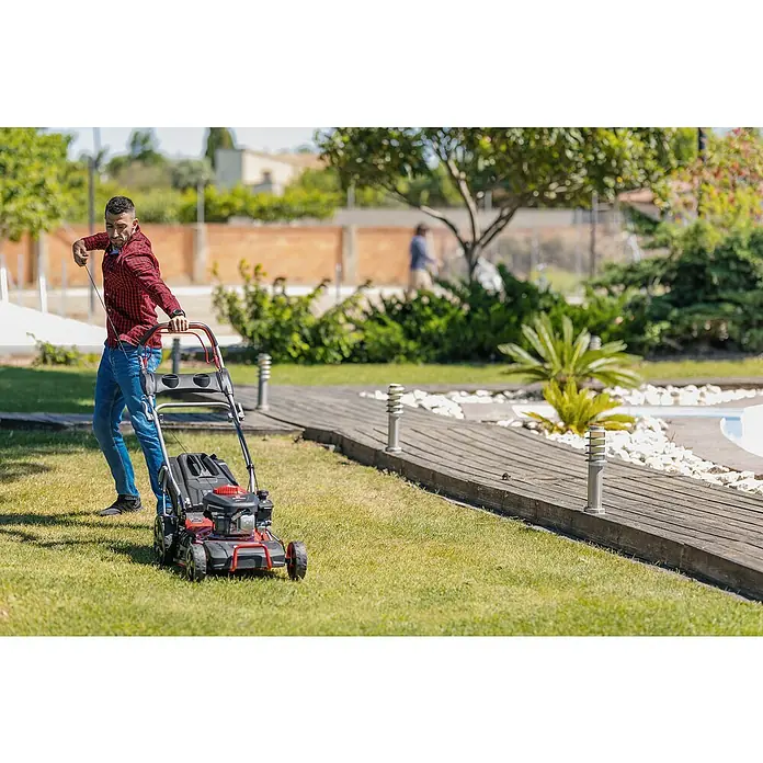 Ducati Cortacésped de gasolina DLM4601 - 2,4 kW, 150 cm³, Ancho de corte: 46 cm - Un hombre tira del cable de arranque de un cortacésped rojo con motor negro, tratando de encenderlo. El césped está bien cuidado y el cielo es azul.