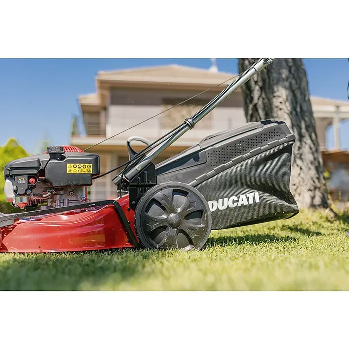 Ducati Cortacésped de gasolina DLM4601 - 2,4 kW, 150 cm³, Ancho de corte: 46 cm - Primer plano de un cortacésped Ducati rojo sobre un césped con una casa al fondo.