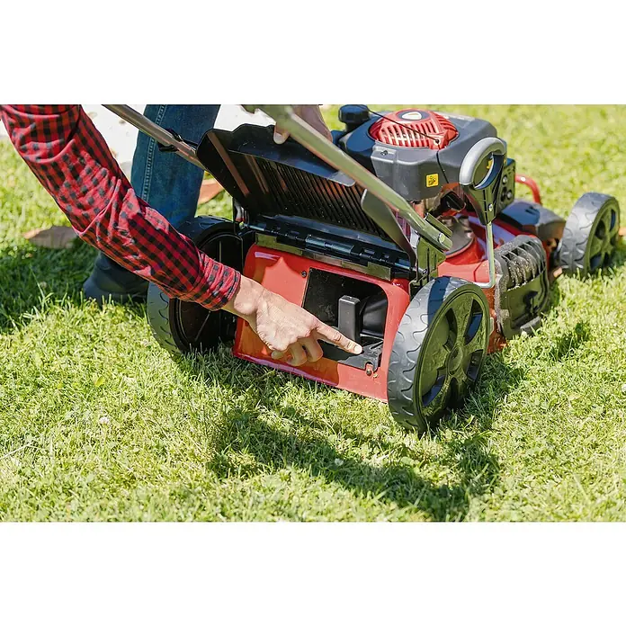 Ducati Cortacésped de gasolina DLM4601 - 2,4 kW, 150 cm³, Ancho de corte: 46 cm - Se inspecciona una cortadora de césped roja. La mano de un hombre con una camisa de cuadros roja y negra señala el dispositivo, que se encuentra en el césped verde.