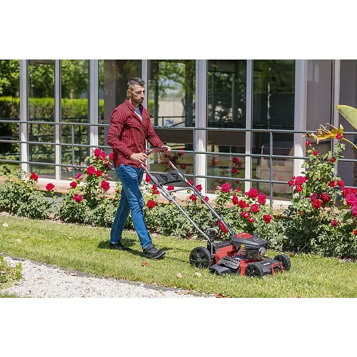 Ducati Cortacésped de gasolina DLM4601 - 2,4 kW, 150 cm³, Ancho de corte: 46 cm - Un hombre corta el césped con una cortadora de césped roja. El producto destaca frente a rosales y un edificio moderno.