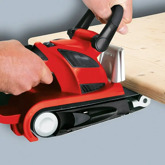 Einhell Expert Lijadora de banda eléctrica TE-BS 8540 E - 850 W - Primer plano de una lijadora de banda roja accionada manualmente sobre una tabla de madera. La herramienta eléctrica se utiliza para lijar.