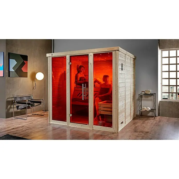 Weka Massivholzsauna Kemi Panorama - Mit 7,5 kW Bio-Aktiv-Ofen mit externer digitaler Steuerung, L x B x H: 210 x 211 x 200 cm, Natur - Innenaufnahme einer Infrarotsauna mit zwei Frauen, die sich entspannen, umgeben von stilvollem Interieur und sanftem, rotem Licht.