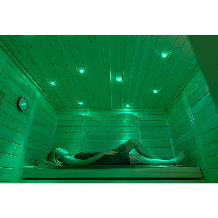 Weka Massivholzsauna Kemi Panorama - Mit 7,5 kW Klassik-Ofen mit externer digitaler Steuerung, L x B x H: 169 x 211 x 200 cm, Natur - Eine entspannte Frau liegt in einer Sauna, beleuchtet von sanftem, grünem Licht, das eine beruhigende Atmosphäre schafft und zur Entspannung einlädt.