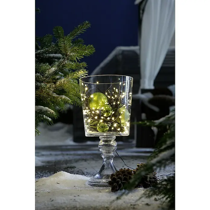 Tween Light Guirnalda luminosa LED - Para exterior, Color de luz: Blanco cálido, Número de bombillas: 120 ud., Funcionamiento en red, 23 m - Una decoración festiva con un jarrón de vidrio sobre un soporte, lleno de adornos verdes y luces, rodeado de nieve y ramas de abeto.