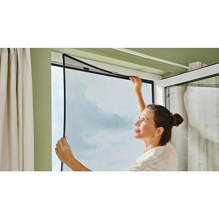 Tesa Insect Stop Mosquitera para ventana - An x Al: 1,2 x 2,4 m, Montaje de la mosquitera: Pinzas, Antracita - Mujer fijando una mosquitera magnética al marco de la ventana. Esta innovadora red protege contra los insectos y asegura aire fresco.
