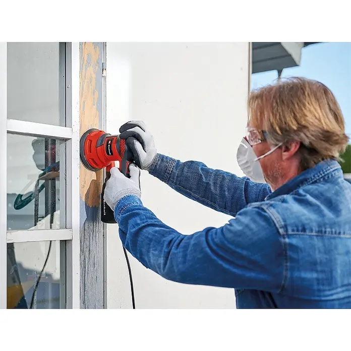 Einhell Classic Lijadora excéntrica eléctrica TC-RS 425 E - 425 W, Diámetro disco de lija: 125 mm - Un hombre lija un marco de ventana con una lijadora orbital Einhell roja, equipado con gafas de seguridad y máscara, para un trabajo seguro y eficaz.