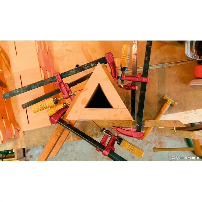 Piher Sargento MM-30 - Anchura de sujeción: 300 mm, Fuerza de tensión: 4.000 N, Apertura máxima: 70 mm - Primer plano de una estructura de madera sujetada con mordazas rojas mientras se seca el pegamento. El diseño es triangular con un agujero en el centro.