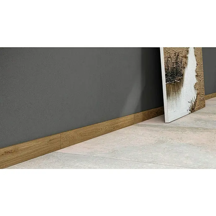 Atlas Concorde Solution Sockelfliese Twist Rovere - 7,2 x 90 cm, Beige, Matt - Nahaufnahme einer braunen Holzfußleiste, die einen grauen Raum von einem cremefarbenen Fliesenboden abgrenzt. Ein Kunstwerk lehnt an der Wand.