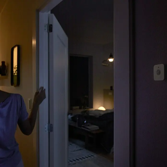 Philips Hue Bewegungsmelder - Weiß, L x B x H: 2 x 5,5 x 5,5 cm - Das Bild zeigt eine Person, die durch eine Tür in ein dunkles Schlafzimmer geht, mit einem Bewegungssensor an der Wand.