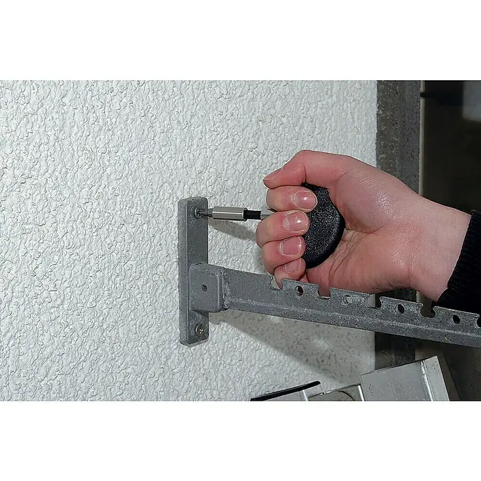 Wolfcraft Hakenschraubendreher - Passend für: Hakenschrauben max. 6 mm Kopfbreite, Ringschrauben max. 6 mm Kopfbreite - Nahaufnahme einer Hand, die mit einem Schraubendreher eine graue Metallhalterung an einer Wand befestigt. Die Halterung hat eine Reihe von Löchern.
