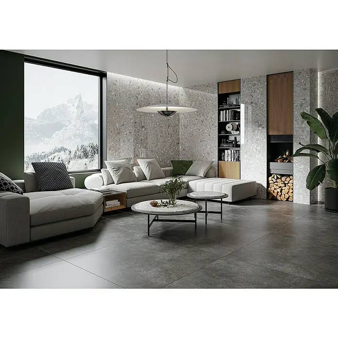 Feinsteinzeugfliese Terrazzo Ceppo - 119,7 x 119,7 cm, Silber, Glänzend - Helles Wohnzimmer mit grauer Couch, Couchtischen, Kamin und Bücherregal. Die grauen Fliesen und die grosse Fensterfront mit Bergblick verleihen dem Raum eine moderne Atmosphäre.
