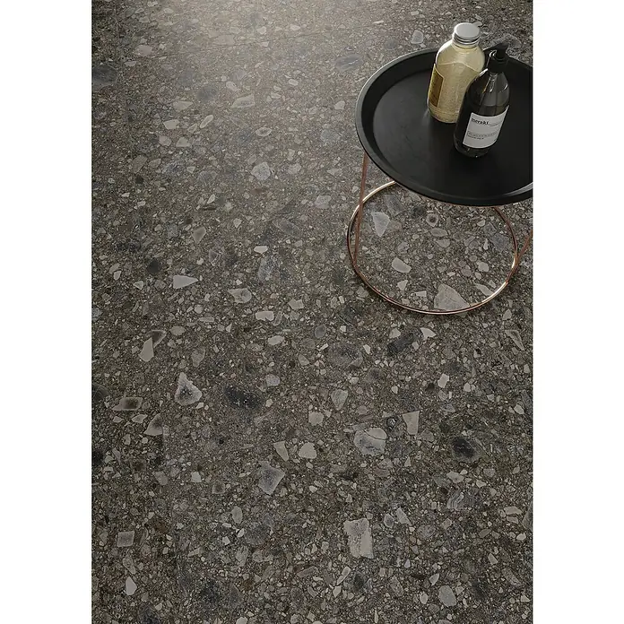 Feinsteinzeugfliese Terrazzo Ceppo119,7 x 119,7 cm, Schwarz, Matt - Elegante graue Terrazzobodenfliese mit kleinen und großen Steinelementen. Daneben steht ein kleiner Beistelltisch mit Flaschen.