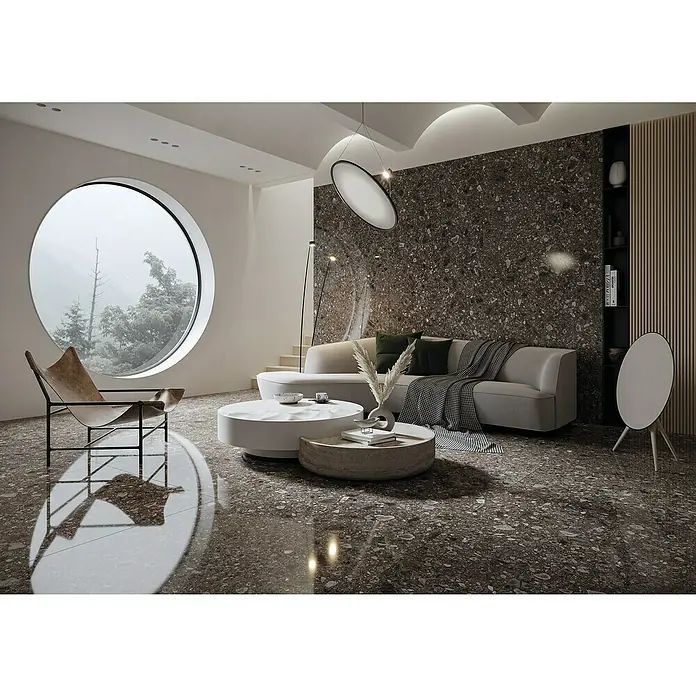 Feinsteinzeugfliese Terrazzo Ceppo - 59,7 x 59,7 cm, Schwarz, Matt - Elegantes Wohnzimmer mit stilvollem Terrazzoboden, modernem Sofa und kreisrundem Fenster, das einen ruhigen Ausblick bietet. Die Beleuchtung akzentuiert das Design.
