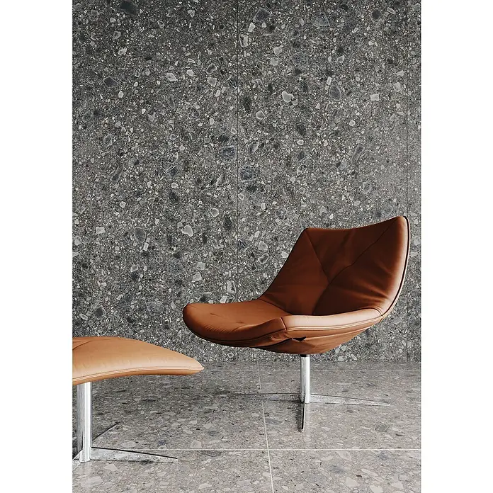 Feinsteinzeugfliese Terrazzo Ceppo - 59,7 x 59,7 cm, Graphit, Glänzend - Ein moderner brauner Ledersessel mit Fußhocker steht vor einer strukturierten Wand. Das Möbelstück hat ein schlankes, zeitgemäßes Design.