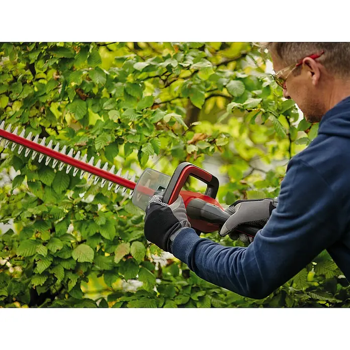 Einhell Power X-Change 18V Akku-Heckenschere GC-CHT 18/45 - 18 V, Li-Ionen, 1 Akku, Schnittlänge: 450 mm - Ein Mann benutzt eine rote Heckenschere, um eine grüne Hecke zu schneiden. Er trägt Schutzbrille und Handschuhe.