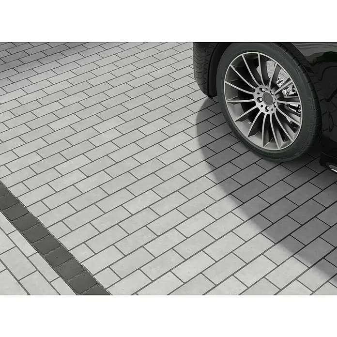 EHL Pflasterstein Drän - Grau, 20 x 10 x 8 cm, Rechteckig - Nahaufnahme eines hellen Pflasterwegs mit einem dunkleren Pflasterstreifen und dem Rad eines Autos, das für eine elegante Einfahrt sorgt.