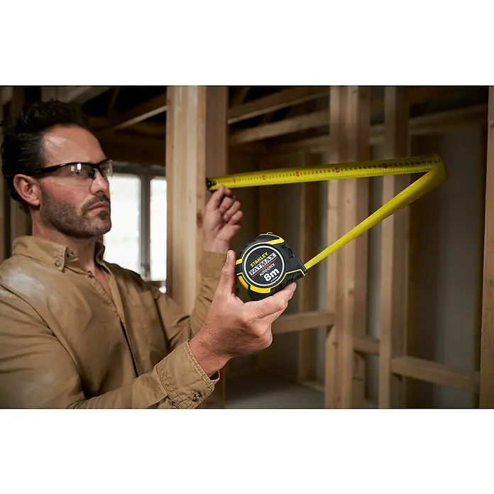 Stanley FatMax Kapselmaßband Autolock - Länge Maßband: 8 m - Ein Mann mit Schutzbrille misst mit einem Stanley Fatmax 8m Rollbandmaß die Ausmaße eines Holzrahmens im Bau.