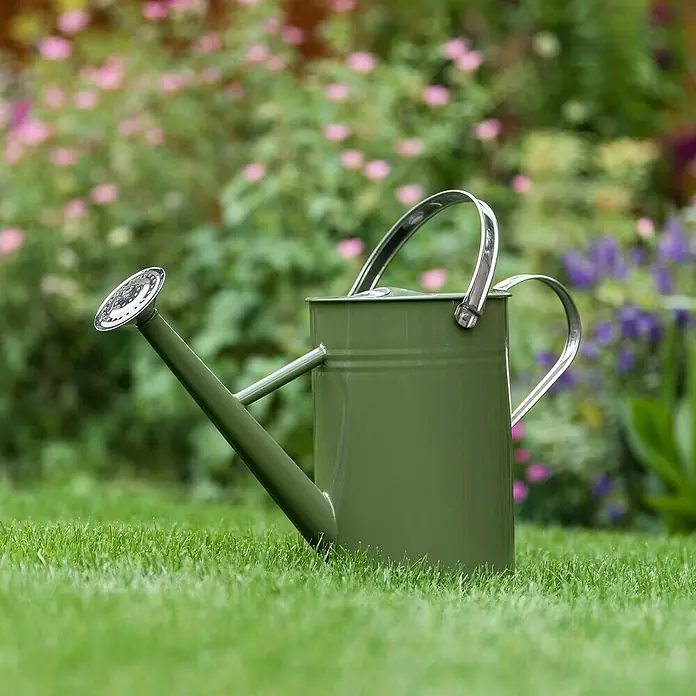 Kent & Stowe Gießkanne - Olivgrün, 4,5 l - Grüne Gießkanne aus Metall mit silberfarbenem Griff und Brausekopf auf Rasen vor blühendem Garten.