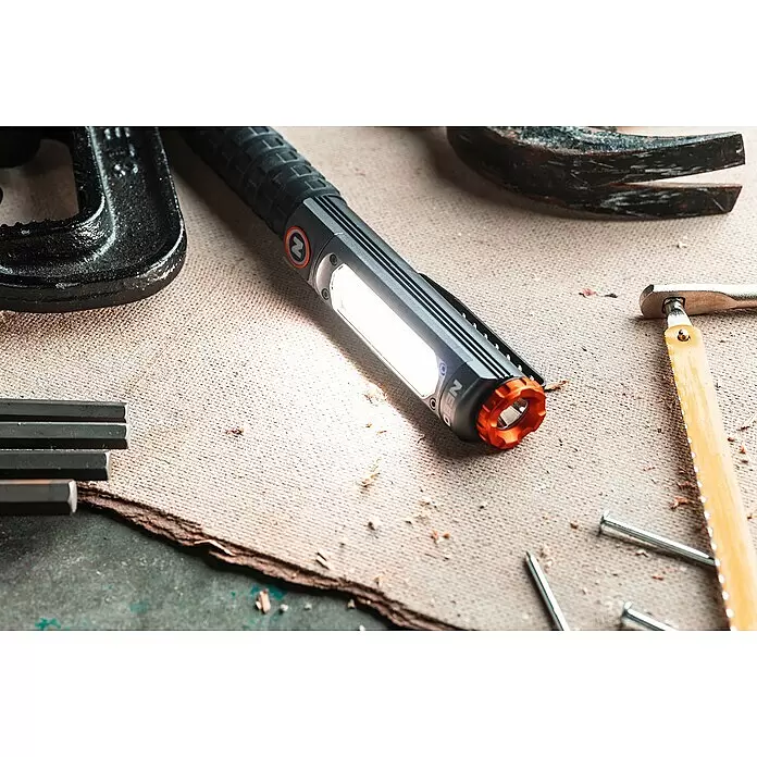 Nebo Tools Linterna Big Larry Pro+ - IPX7 - Linterna NEBO negra y naranja iluminada, entre herramientas y virutas de madera en un banco de trabajo.