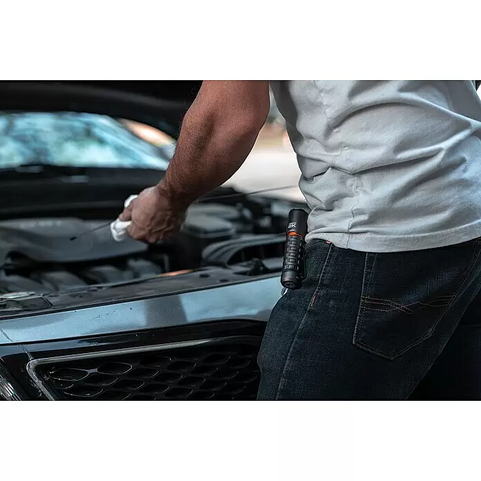 Nebo Tools Linterna Torchy 2K - 2.000 lm - Linterna Torchy 2K negra y naranja enganchada a los vaqueros durante el mantenimiento del coche.