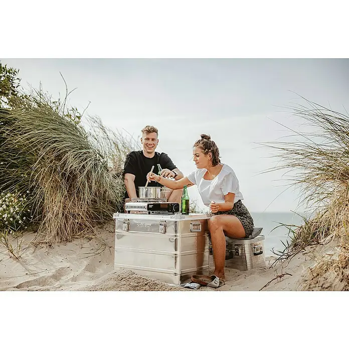 Enders Campingkocher Maveriq - 2,2 kW - Kochkiste von Enders im Einsatz am Strand mit zwei Personen, die kochen und Bier trinken.
