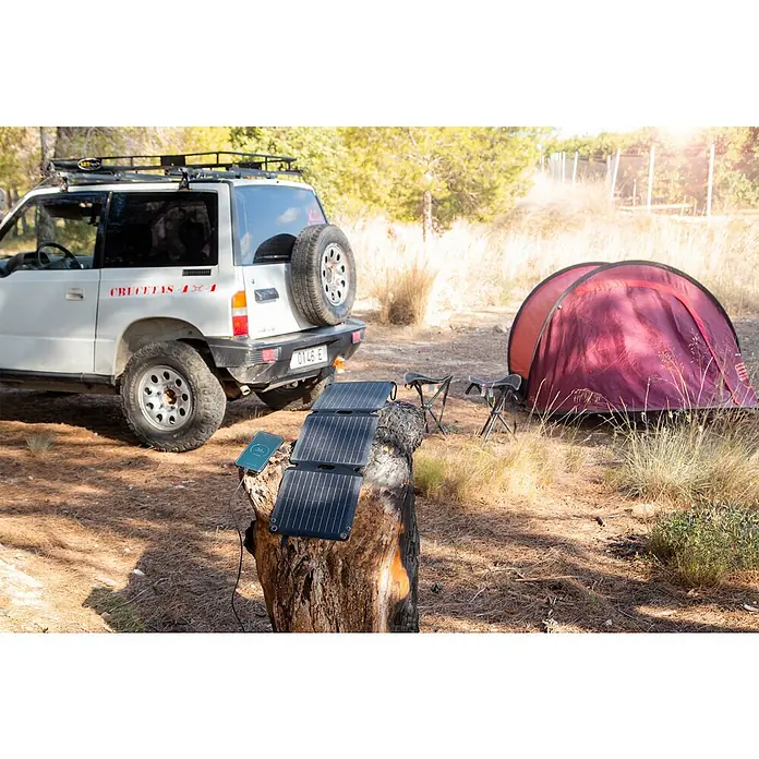 Panel solar plegable Innovate - 21 W, 2 conexiones USB - Escena de camping con un panel solar portátil cargando un smartphone, con un todoterreno y una tienda de campaña al fondo.