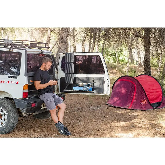 Panel solar plegable Innovate - 21 W, 2 conexiones USB - Un hombre se apoya en un SUV blanco con una cocina integrada mientras usa su móvil. Al fondo, hay una tienda roja.