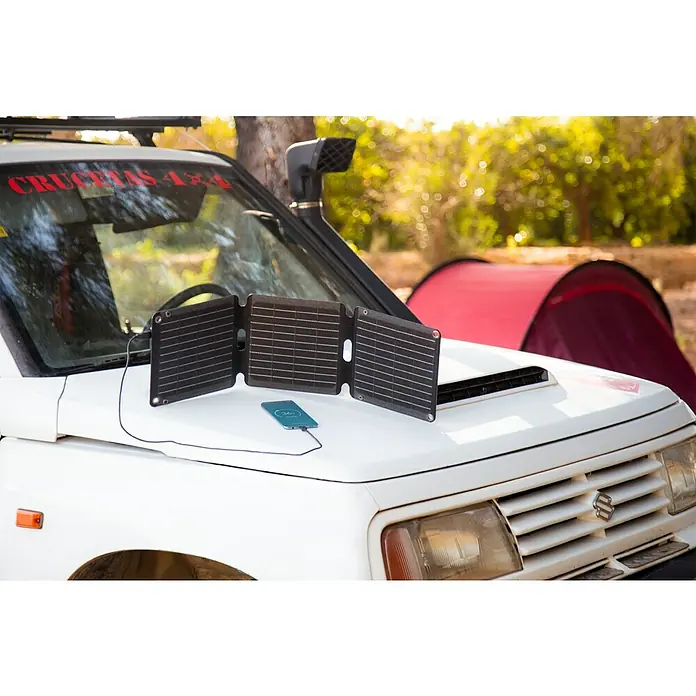 Panel solar plegable Innovate - 21 W, 2 conexiones USB - Un panel solar portátil carga un teléfono inteligente en el capó de un SUV blanco, con letras rojas al fondo de un camping.