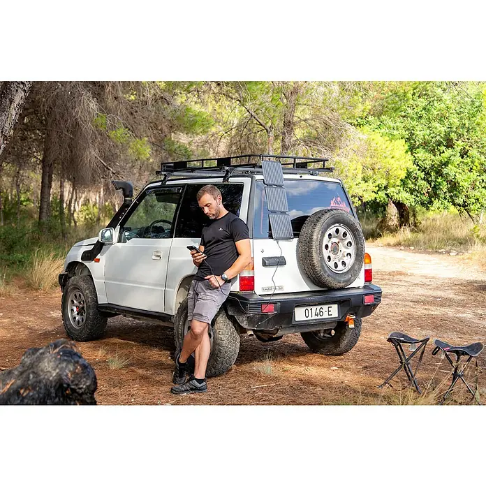 Panel solar plegable Innovate - 21 W, 2 conexiones USB - Hombre con panel solar conectado al coche. El panel está cargando su teléfono mientras está al aire libre. También se ve material de acampada.
