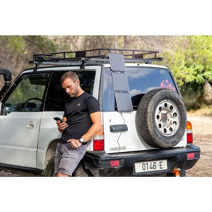Panel solar plegable Innovate - 21 W, 2 conexiones USB - Un hombre se apoya en un SUV y utiliza un cargador solar para alimentar su teléfono. El panel solar está fijado al vehículo.