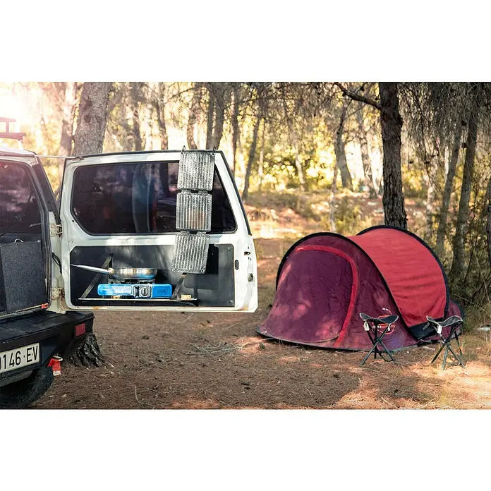 Panel solar plegable Innovate - 21 W, 2 conexiones USB - Escena de camping con la puerta de un coche sirviendo como cocina, con hornillo y tienda, ideal para aventuras al aire libre y comidas.