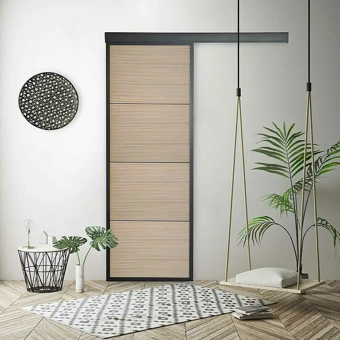 Kit atelier Holzschiebetür Loft Holz District - 930 x 2.128 mm, Schwarz Sandstruktur, Eichenholznachbildung - Ein modernes Interieur mit einer hellbraunen Schiebetür mit schwarzen Akzenten, ergänzt durch einen stilvollen Teppich und eine Hängematte.
