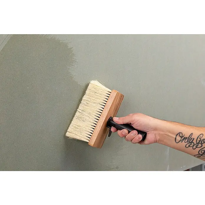 Knauf Tiefengrund - 5 l - Eine Hand hält eine Tapezierbürste an eine grüne Wand, wobei die Farbe der Wand durch das Werkzeug verändert wird.