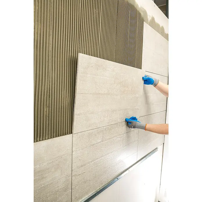 Knauf Flexkleber Extra - 20 kg - Anbringen einer grauen Wandfliese an einer Wand mit Kleber, wobei blaue Handschuhe für eine sichere Installation sorgen.