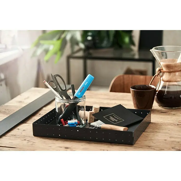 Bosch Akku-Heißklebestift Gluey - Farbe: Smoky Grey - Tischszene mit einer schwarzen Ablage, gefüllt mit Büroutensilien wie Stiften, einer Schere und einem Bosch-Klebestift, neben einer Kaffeekanne.