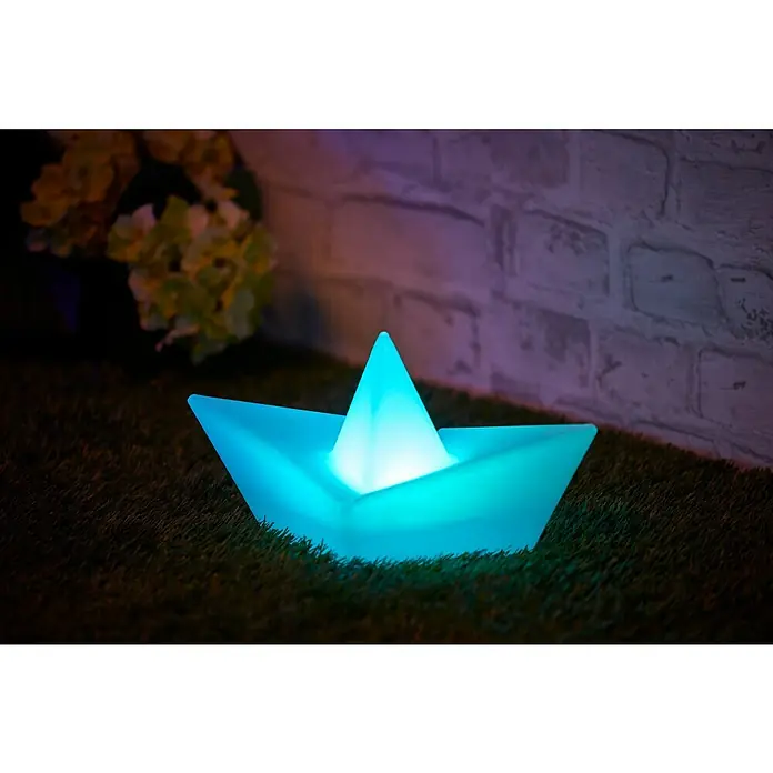 BAUHAUS Solar-Dekoleuchte Schiff - Weiß, IP65 - Leuchtendes Papierboot auf Rasen mit Ziegelmauer und Blumen im Hintergrund. Ein dekoratives Highlight für jeden Raum.