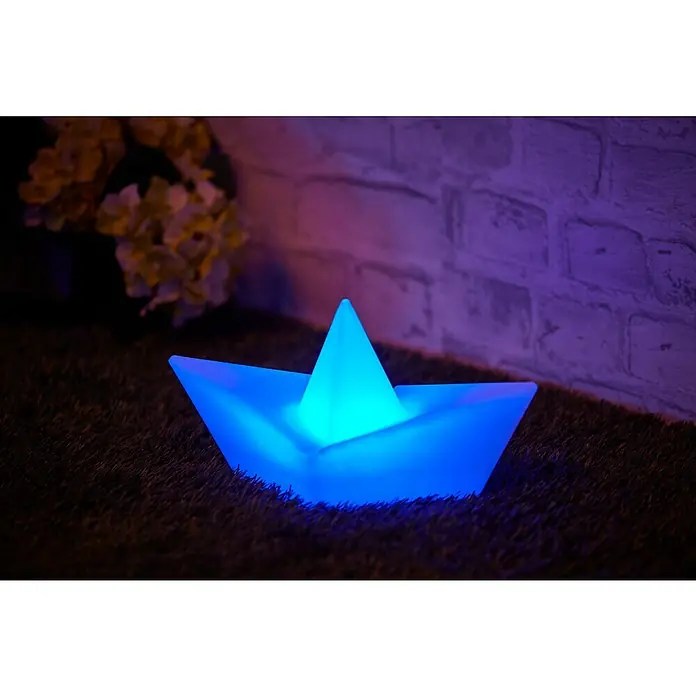 BAUHAUS Solar-Dekoleuchte Schiff - Weiß, IP65 - Nahaufnahme eines leuchtenden, blauen Papierboots auf Gras mit Blumen und einer Backsteinmauer im Hintergrund.